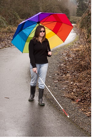 a blind woman using a cane to navigate a paved wooded path, she is carrying a rainbow umbrella